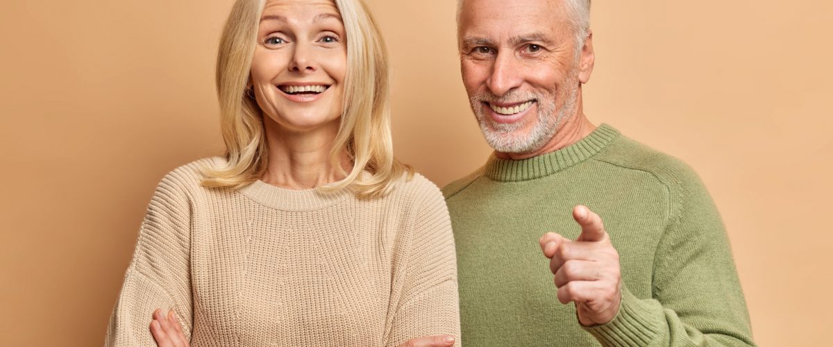 Portrait of happy aged woman and man stand closely to each other dressed in casual jumpers look with great interest in front isolated over brown studio background. People age and emotions concept Portrait of happy aged woman and man stand closely to each other dressed in casual jumpers look with great interest in front isolated over brown studio background. People age and emotions concept